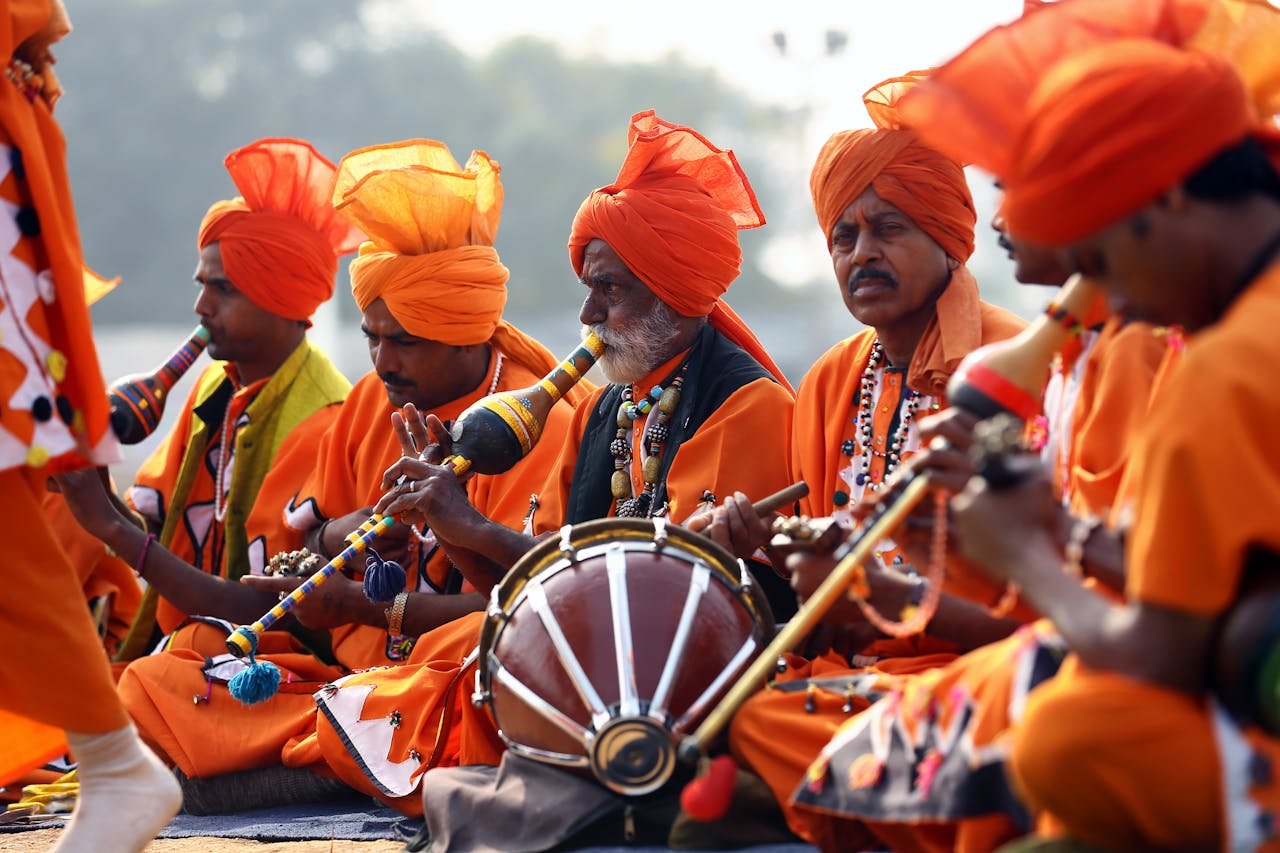 hero-img-01 A group of Indian men in vibrant orange traditional attire performing music during a cultural festival outdoors.